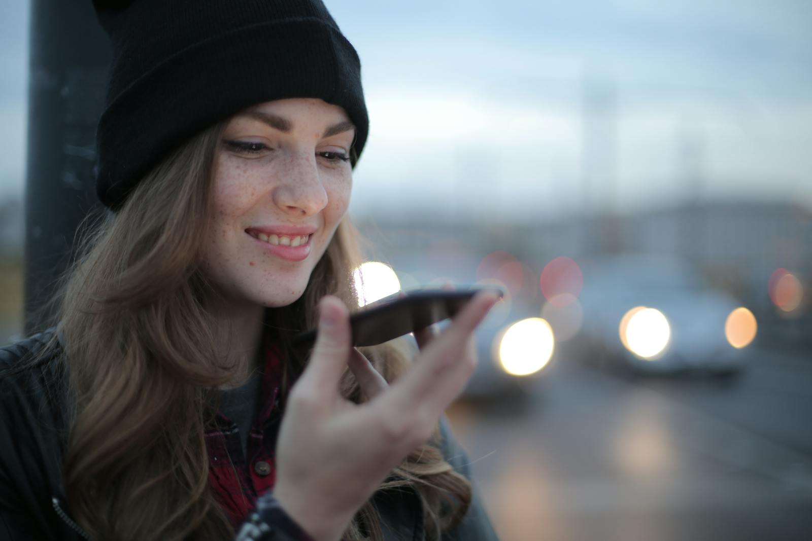 Young woman smiling while using a smartphone on an urban street during twilight.